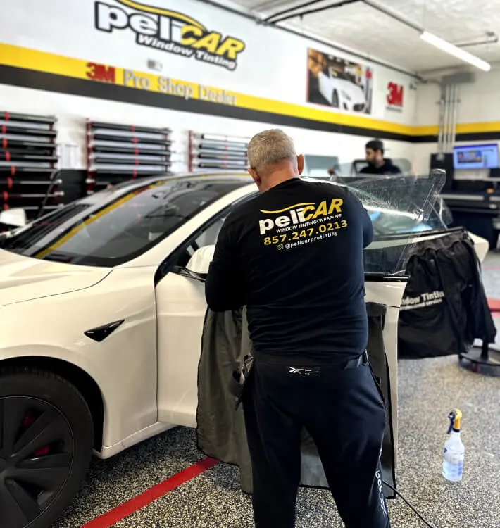 A professional window tinter applying a tint film to the front window of a white Tesla at peliCAR Window Tinting. The technician, wearing a branded peliCAR shirt, is carefully smoothing out the film while the shop’s equipment and branding are visible in the background.