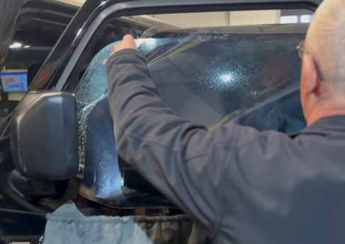 A professional window tinter applying a dark tint film to the front window of a black vehicle. The installer, seen from behind, carefully positions the film while using a cloth to protect the door panel.