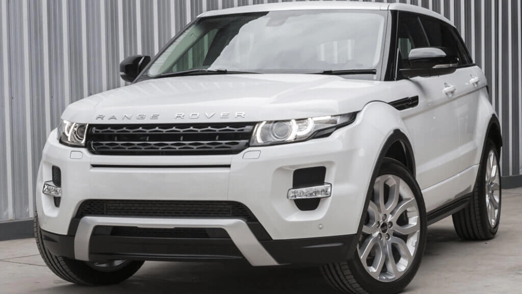 A white Range Rover Evoque parked in front of a corrugated metal wall. The luxury compact SUV has a bold front grille, sleek LED headlights, and large silver alloy wheels. The vehicle’s glossy paint reflects the surroundings, highlighting its clean and polished exterior.