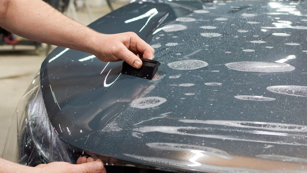 A close-up of a person using a squeegee to smooth out protective film on a car's hood, with water droplets visible on the surface.