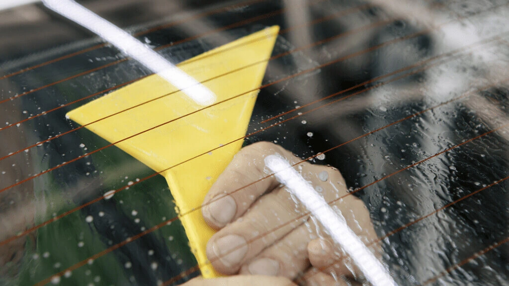 A hand holding a yellow squeegee tool on the back window of a vehicle with visible water droplets on the glass.