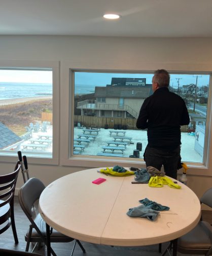 A professional window tinter installing film on a large window inside a beachfront property. The room has round tables with cleaning supplies, cloths, and spray bottles, while the view outside shows picnic tables, a sandy beach, and ocean waves.