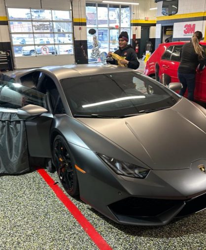 A matte gray Lamborghini Huracán in a professional tinting shop, with a technician installing window tint on the driver’s side. The car features black wheels, orange brake calipers, and an aggressive design. In the background, two additional technicians are working on a red vehicle, with 3M branding visible on the walls.