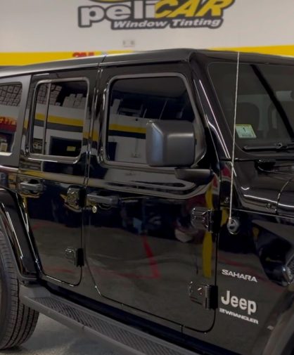 A black Jeep Wrangler Sahara parked inside a professional window tinting shop. The vehicle features tinted windows, rugged off-road styling, and chrome accents. The "peliCAR Window Tinting" branding is visible on the wall in the background.