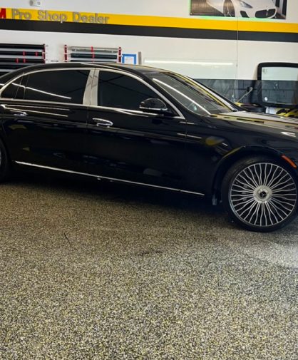 A black Mercedes-Maybach luxury sedan parked inside a professional window tinting shop. The car features tinted windows, chrome accents, and distinctive multi-spoke wheels. The "peliCAR Window Tinting" branding and 3M Pro Shop Dealer signage are visible on the wall in the background.