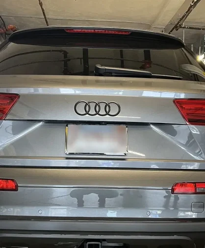 A rear view of a silver Audi Q7 3.0T parked inside a professional window tinting shop. The SUV features tinted windows, sleek LED taillights, and a polished exterior. The "peliCAR Window Tinting" branding is reflected in the vehicle's surface, and various tools and tinting supplies are visible in the background.