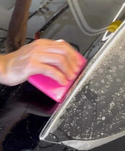 A close-up of a person applying paint protection film on a black car using a pink squeegee.