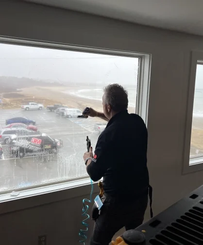 A person cleaning a large window with a squeegee while looking out at a beach scene from the window.