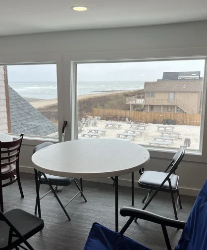 A room with two large windows overlooking the beach, a table and chairs are set up in the room.