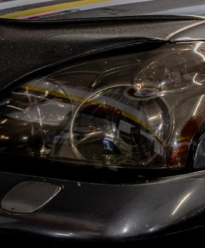 Close-up of a car's headlight on a dark-colored vehicle, reflecting the surrounding workshop environment. The lens appears clean and well-maintained, with reflections of overhead lights and signage visible. The hood and fender have a slight sheen, indicating recent cleaning or detailing.