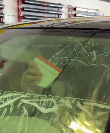 A close-up of a professional installing window tint on a car's windshield using a squeegee. The tint film is being smoothed out over the glass, with soapy water and air bubbles visible beneath the surface. In the background, rolls of window tint film are mounted on racks in a professional tinting shop.