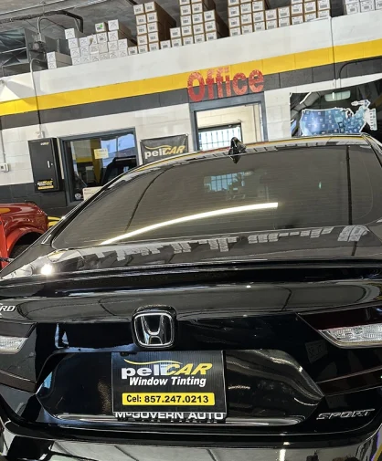 A black Honda Accord Sport with freshly tinted windows parked inside a professional tinting shop. The license plate frame displays the Pelicar Window Tinting logo and contact number. The background features an 'Office' sign, stacked tint film boxes, and a red truck with its door open, indicating an active workshop.